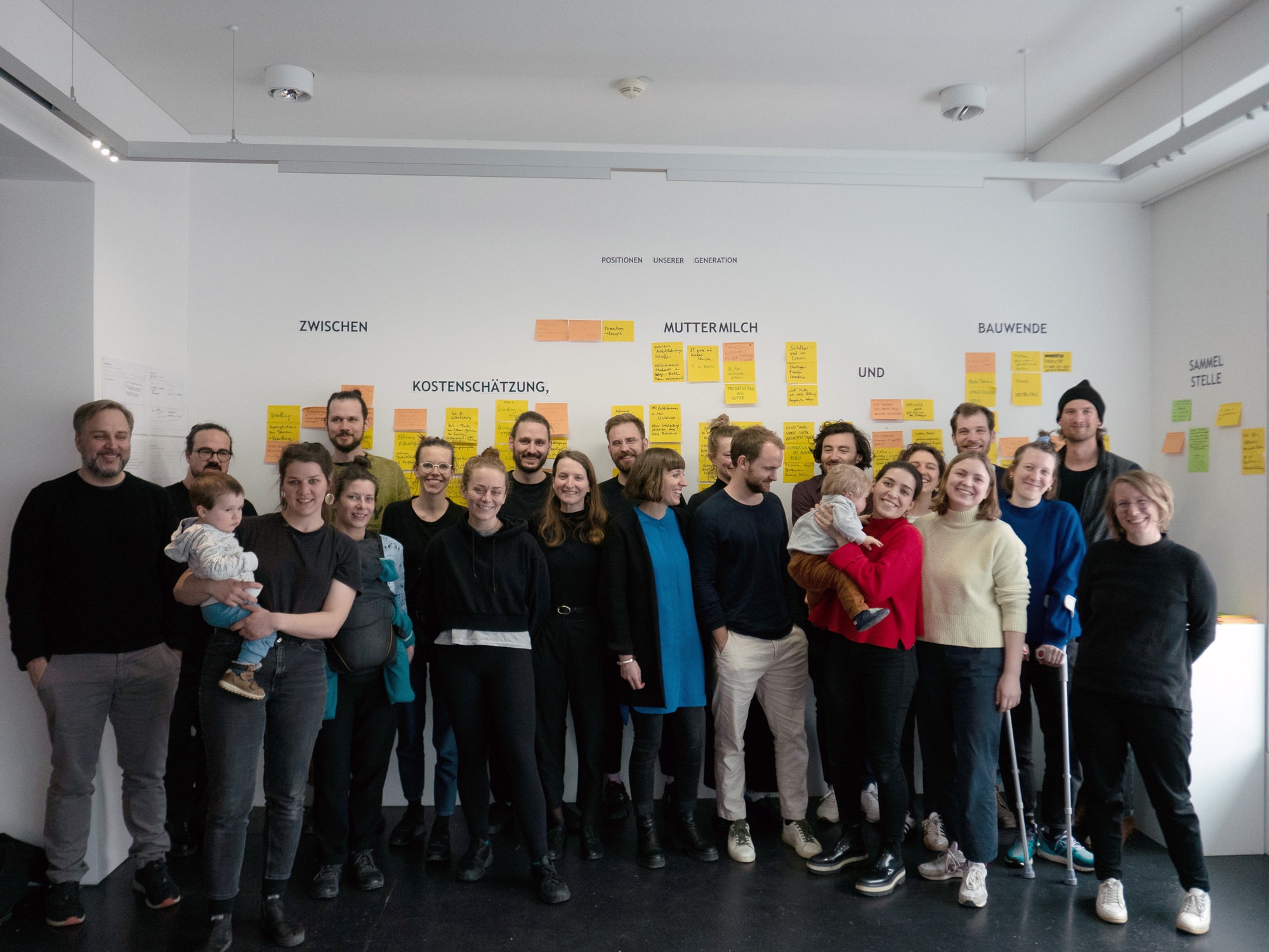 Group photo of workshop participants, some holding small children, against a metal wall on a sunny terrace asphalt kollektiv