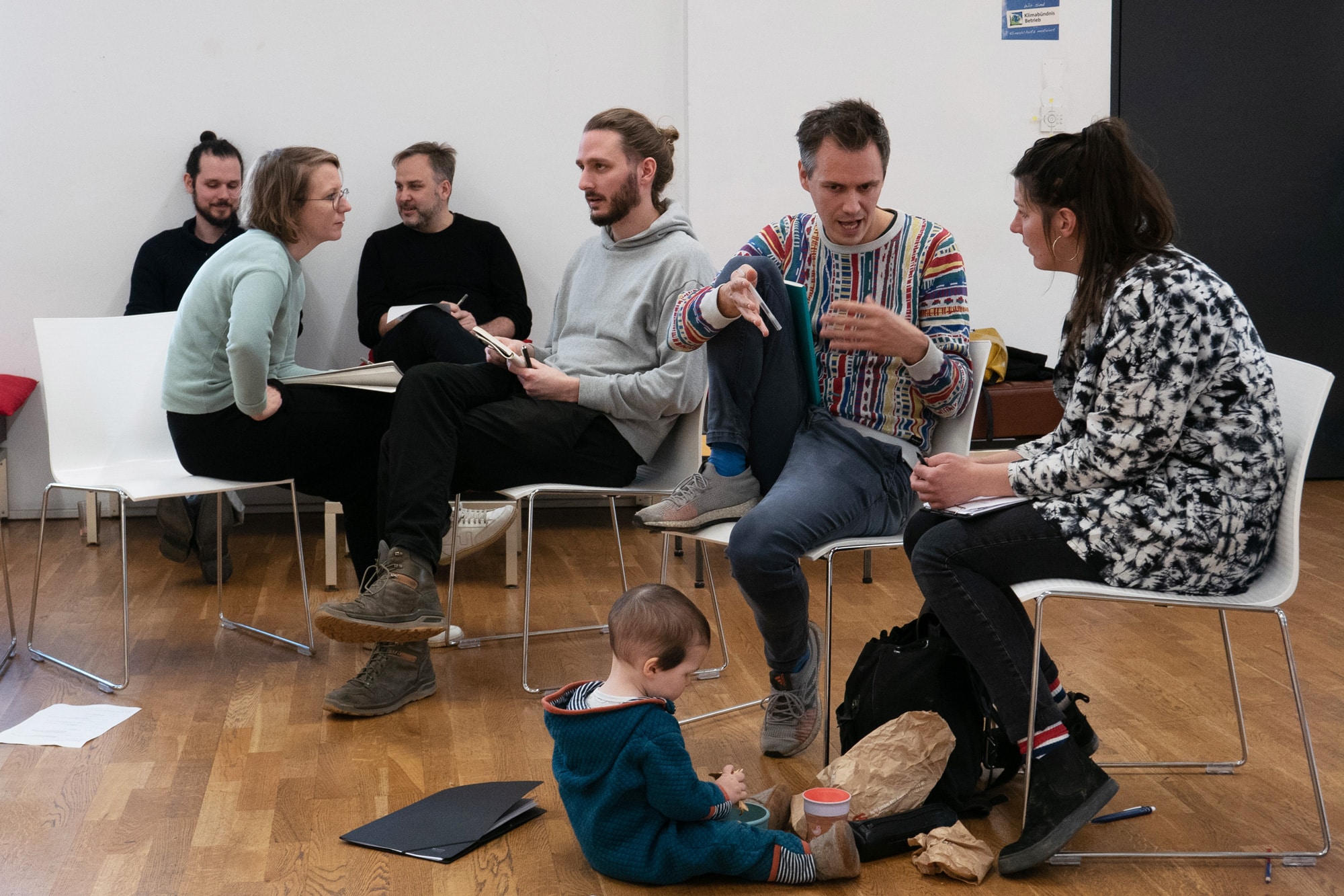 Small group talking on chairs in a circle while a toddler plays on the floor asphalt kollektiv